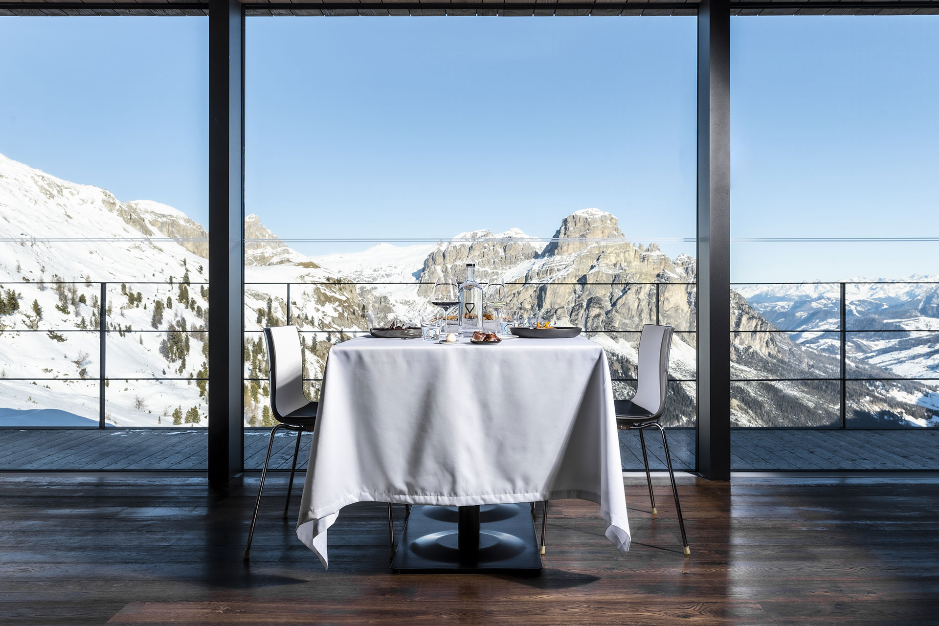 Vista dall'interno del KELINA Restaurant, dove sono posti file di tavoli bianchi, sedie e divanetti. Su un bordo è posta in esposizione una serie di vini differenti.