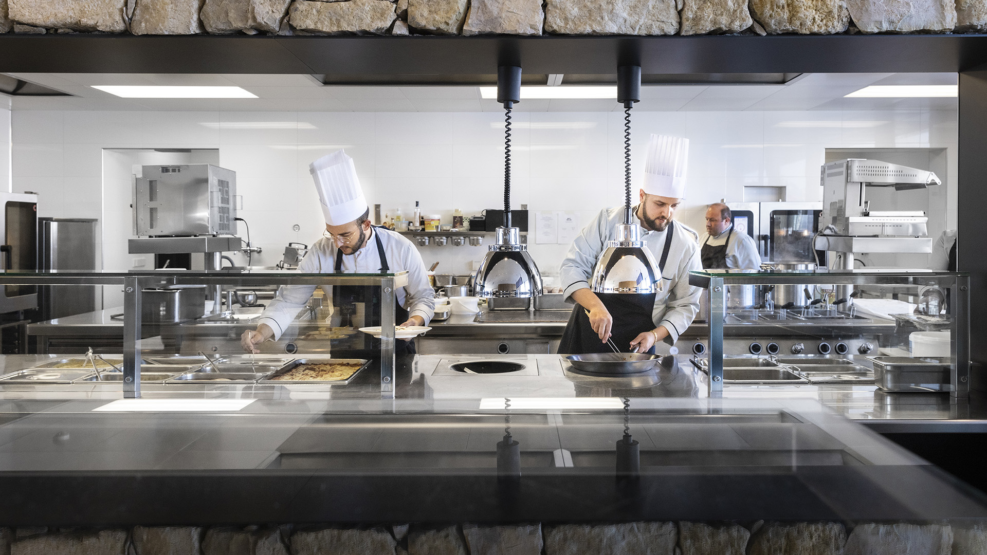 Prospettiva frontale della cucina del Piz Boè dove tre cuochi stanno preparando il pranzo da fornire ai clienti nell'area self-service.
